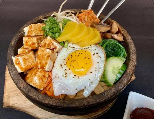 A stone bowl filled with rice, vegetables, tofu, a fried egg, and garnishes, served with a side of sauce.