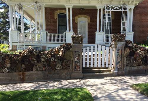 A decorative stone wall with unique textures and patterns surrounds a white picket gate in front of a historic house.