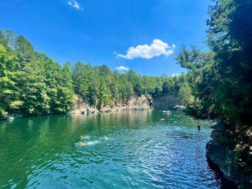 A serene lake surrounded by lush green trees, with people swimming and a clear blue sky above.