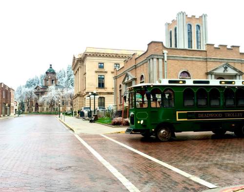 A green trolley bus turns onto a brick street, with historic buildings and frosted trees in the background.