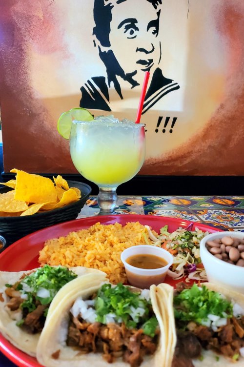 A vibrant plate of tacos with rice, salsa, and a margarita, set against a colorful mural and tortilla chips.