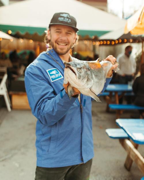 A smiling man in a blue jacket holds a large fish, standing outdoors at a lively market or festival.