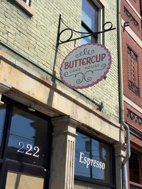 Sign for "Buttercup Cake House" hanging above a storefront, with "Espresso" visible in the window below.
