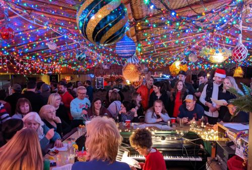 A lively bar scene decorated with colorful lights, featuring a piano, festive ornaments, and a crowd enjoying the atmosphere.