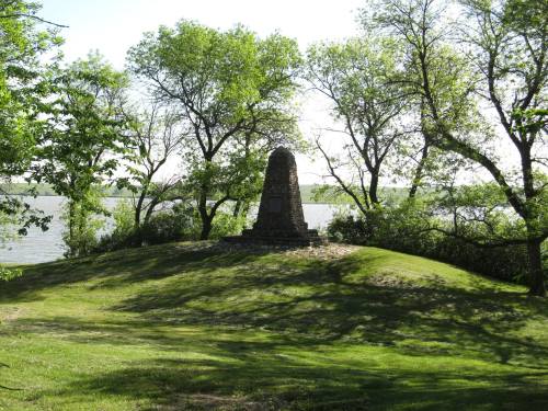 A stone monument on a grassy hill surrounded by trees, overlooking a calm body of water under a clear sky.