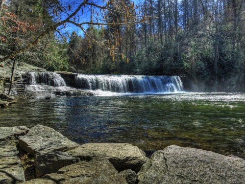 A serene waterfall cascades over rocks into a clear pool, surrounded by lush greenery and trees under a blue sky.
