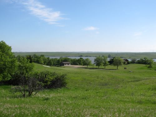 A serene landscape featuring green hills, a small building, and a calm lake under a clear blue sky.
