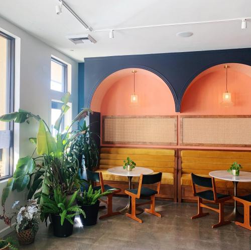A cozy café interior with yellow seating, round tables, and lush plants, featuring arched walls and warm lighting.