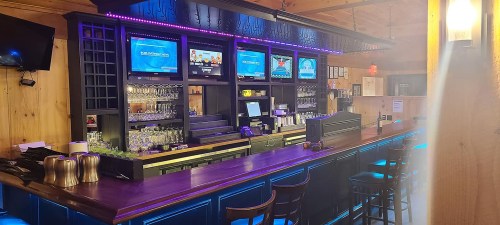 A modern bar with a wooden interior, featuring multiple screens, a well-stocked shelf, and illuminated counter.