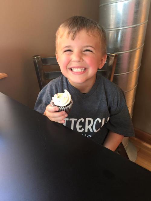 A smiling young boy in a gray shirt holds a cupcake while sitting at a table.
