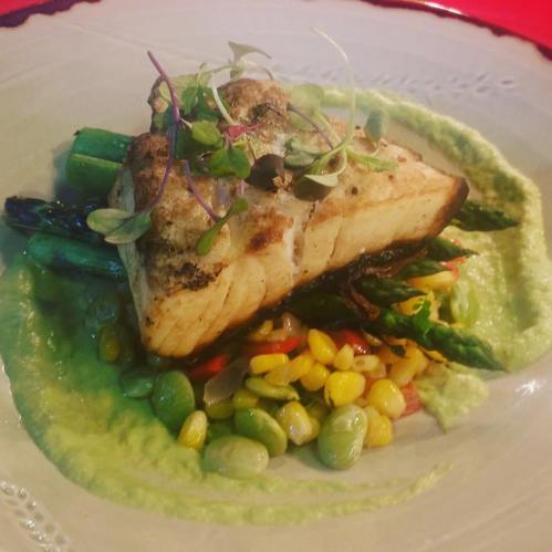 A beautifully plated dish featuring grilled fish atop asparagus and colorful vegetables, with a green puree on the side.