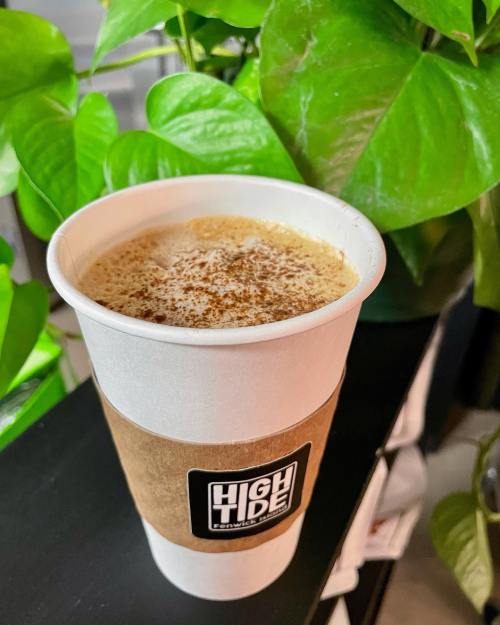 A paper coffee cup with a "High Tide" logo, filled with frothy coffee, surrounded by green plants.