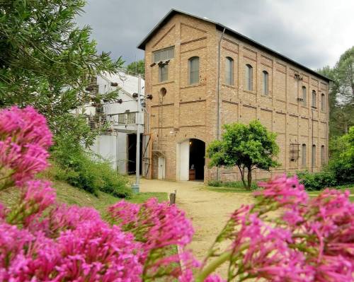 A brick building surrounded by greenery and pink flowers, under a cloudy sky.