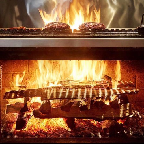 A grill with two burgers cooking over a vibrant fire, with flames and glowing embers visible below.