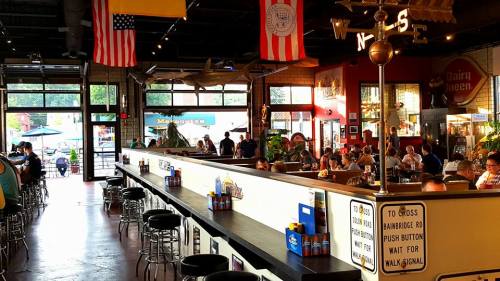 A lively restaurant interior with patrons dining, flags hanging, and a long bar area filled with drinks.