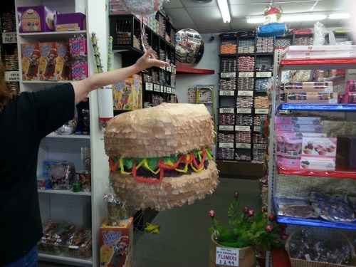 A person holds a colorful hamburger-shaped piñata in a candy store filled with various treats and decorations.