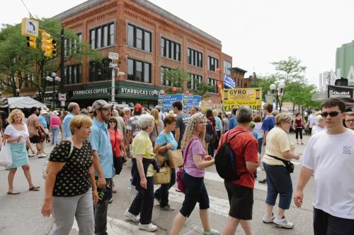 A diverse crowd of people walking in a street festival, with banners and food stalls in the background.