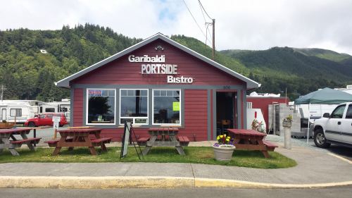 A red bistro building with "Garibaldi PORTSIDE Bistro" sign, picnic tables outside, surrounded by greenery and mountains.