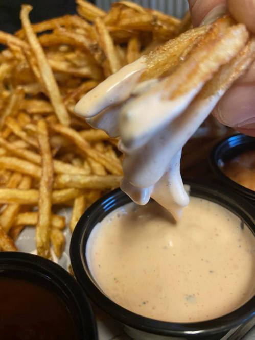 A hand dips a crispy French fry into a creamy sauce, with more fries and sauce containers in the background.