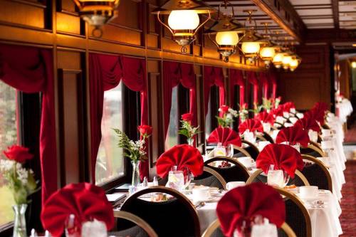 Elegant dining car with tables set for a meal, featuring red napkins and roses, illuminated by vintage lamps.