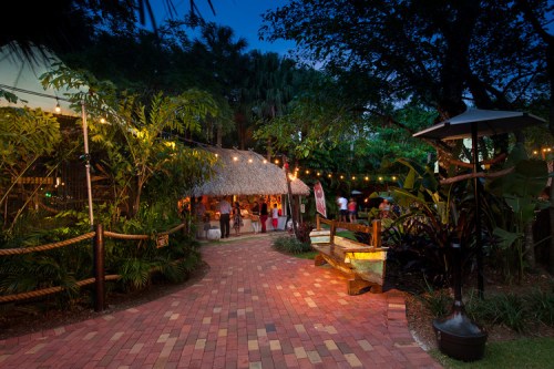 A brick pathway leads to a thatched-roof building, surrounded by lush greenery and string lights at dusk.