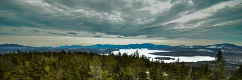 A panoramic view of a lake surrounded by mountains and dense forests under a cloudy sky.