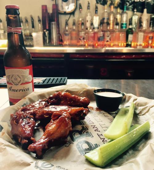 A plate of saucy chicken wings with celery sticks and a dipping sauce, alongside a bottle of beer, in a bar setting.