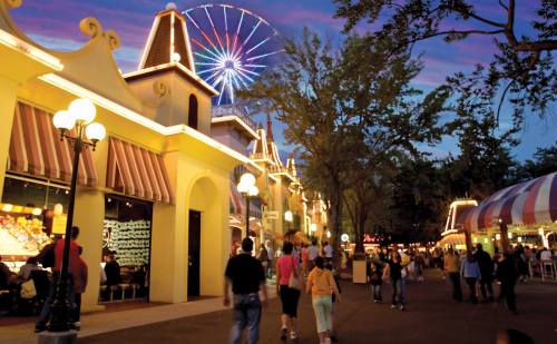 A vibrant amusement park scene at dusk, featuring colorful buildings, a Ferris wheel, and people strolling along the path.