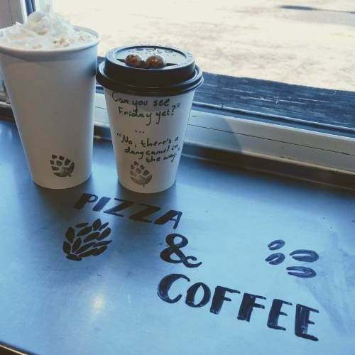 Two coffee cups on a table with a humorous quote, alongside the words "PIZZA & COFFEE" painted on the surface.