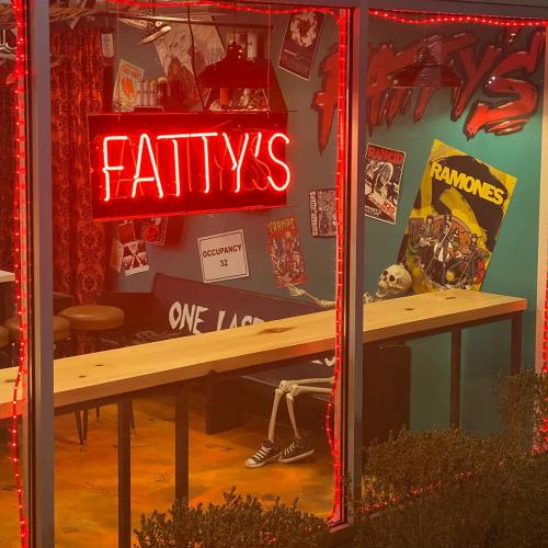 A neon sign reads "FATTY'S" in a colorful bar with a skeleton sitting at a table, surrounded by punk rock posters.