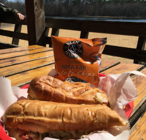 Two sandwiches on a red plate with a bag of potato chips, set against a scenic outdoor view by a lake.