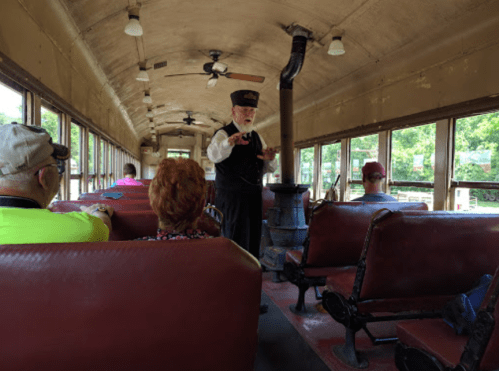 A conductor in uniform speaks to passengers inside a vintage train car with wooden benches and a stove.