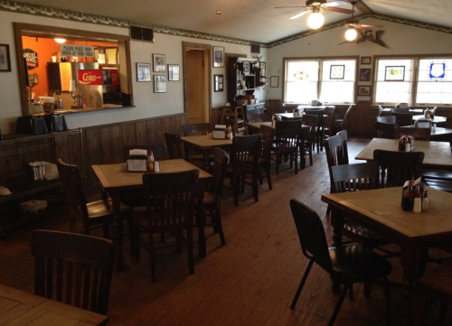 Cozy restaurant interior with wooden tables and chairs, featuring a counter and framed pictures on the walls.