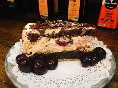 A slice of layered chocolate cake with cherries, cream, and chocolate shavings, served on a decorative plate.