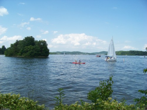 A serene lake scene with a kayak and a sailboat, surrounded by green trees and a clear blue sky.