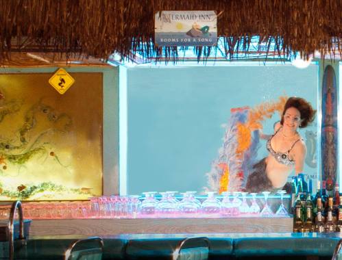 A woman in a mermaid costume swims near a bar with tropical decor and colorful drinks.