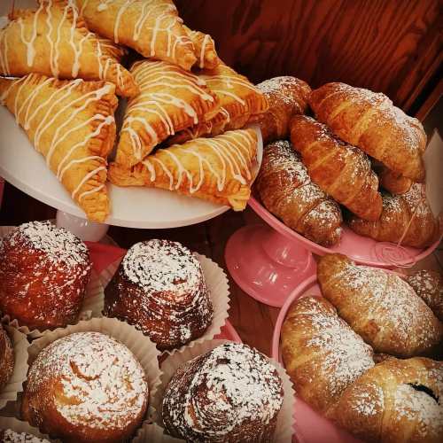 A display of assorted pastries, including drizzled croissants and powdered sugar-dusted muffins on pink stands.