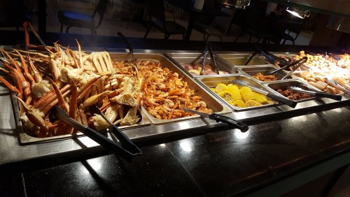A seafood buffet featuring crab legs, shrimp, corn, and various other dishes in metal trays.