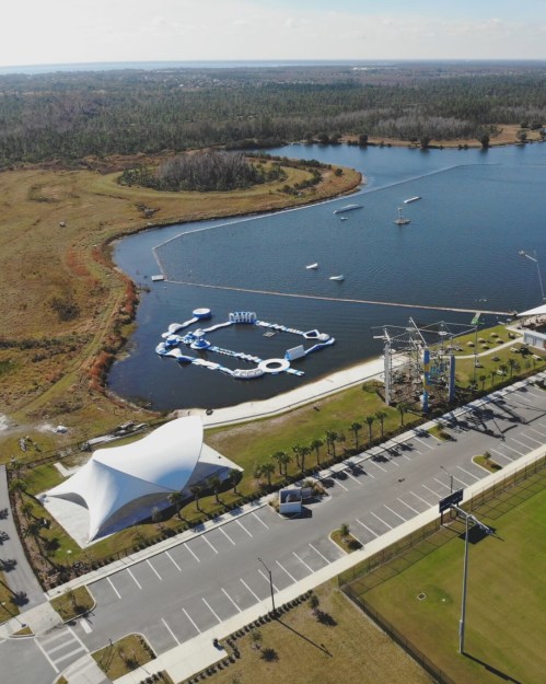 Aerial view of a lake with inflatable water park features, surrounded by greenery and a parking area.