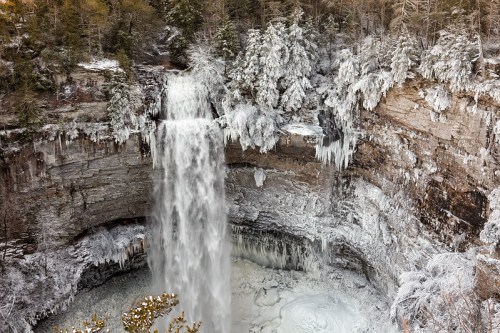 A frozen waterfall cascades into a snowy, icy basin, surrounded by frosted trees and rocky cliffs.