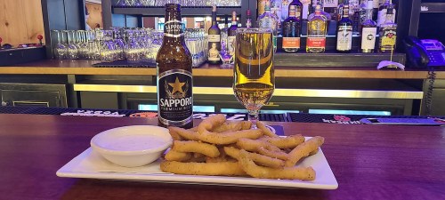 A plate of crispy fried food with a dipping sauce, alongside a beer bottle and a glass of beer on a bar counter.