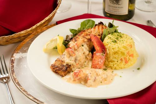A plate of grilled salmon with creamy sauce, lobster, rice, and a side salad, garnished with lemon and herbs.