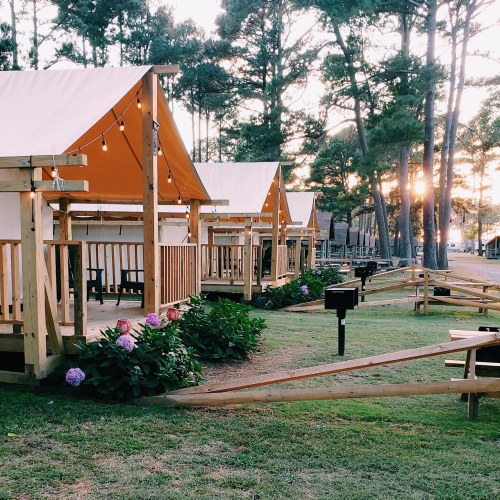 Cozy glamping tents with wooden frames and string lights, surrounded by flowers and trees at sunset.