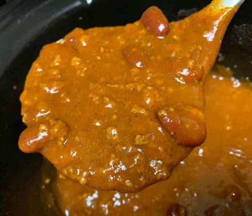 A close-up of a spoonful of thick chili with kidney beans, lifted from a pot.
