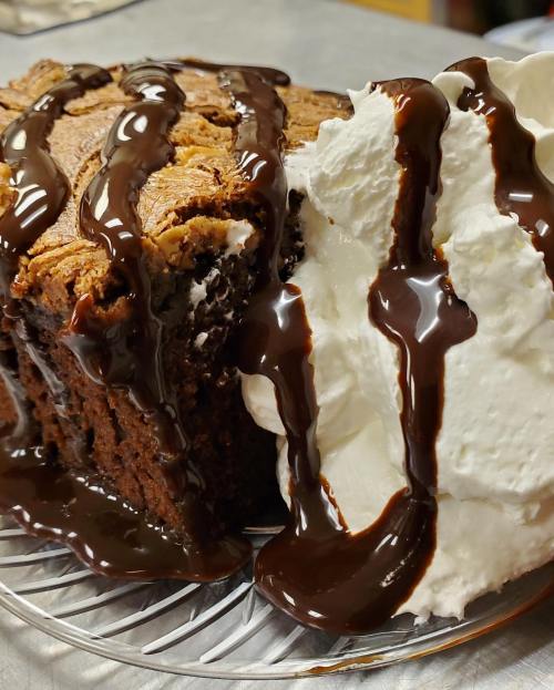 A slice of rich chocolate cake topped with whipped cream and drizzled with chocolate sauce on a glass plate.