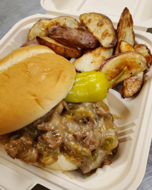 A sandwich with beef, melted cheese, and a pepper, served with crispy potato wedges in a takeout container.
