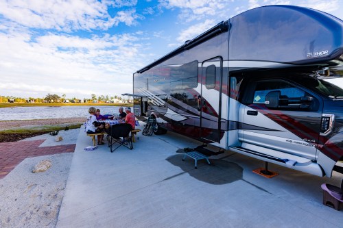 A group of people enjoying a meal outside an RV by a waterfront, with a clear blue sky and scenic surroundings.
