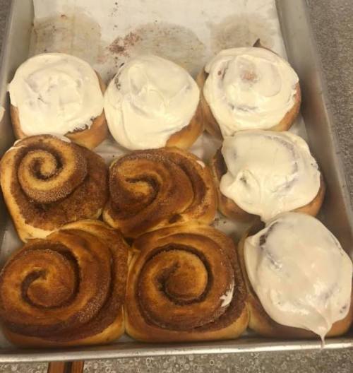 A tray of freshly baked cinnamon rolls, some topped with cream cheese frosting, arranged in two rows.