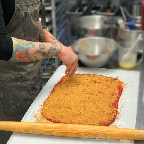 A person with tattoos spreads a layer of brown sugar on a red dough in a kitchen setting. A rolling pin is nearby.
