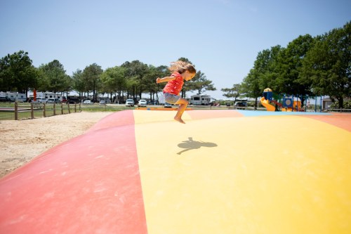 A child jumps joyfully on a large inflatable bounce pad at a sunny playground. Trees and campers are in the background.